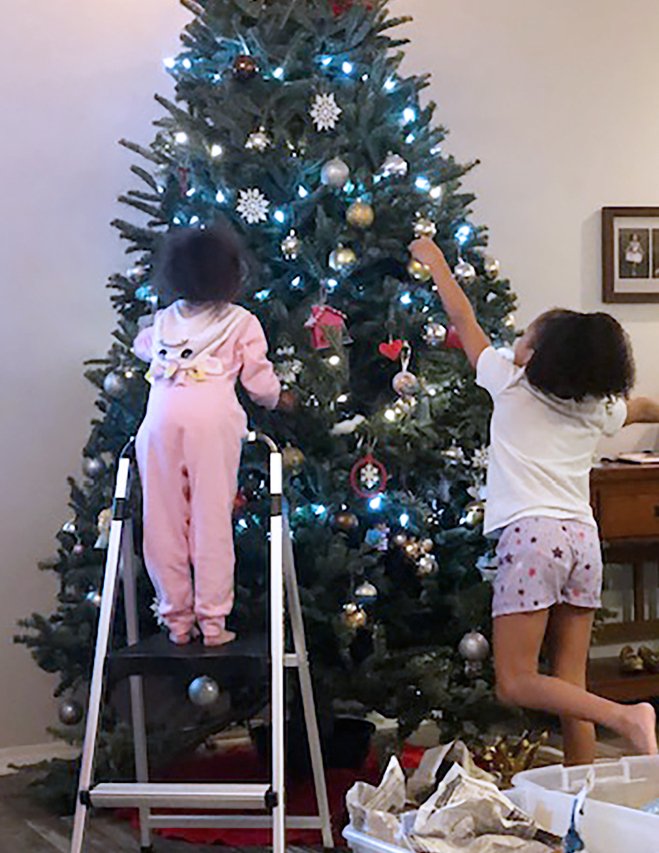 Sisters decorating a Christmas tree together, one reaching up on a ladder while the other hangs ornaments below