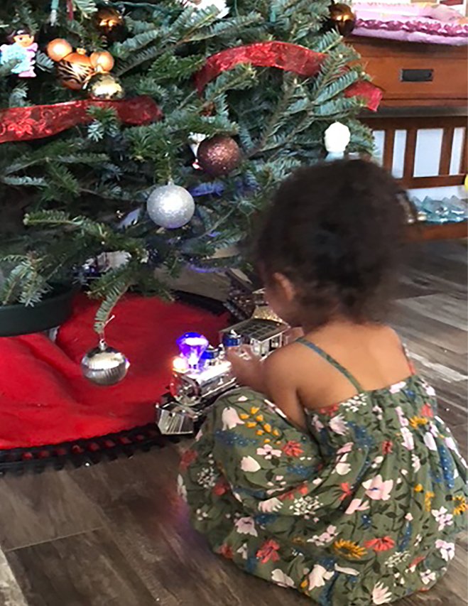 Young girl sitting by the Christmas tree, mesmerized as a toy train circles beneath the glowing lights.